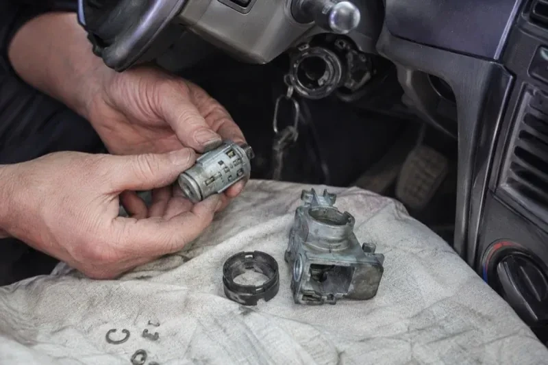 Hands holding a removed ignition cylinder beside a car steering column, with the housing and small parts on a cloth. — Ignition Lock Repair, St. Catharines